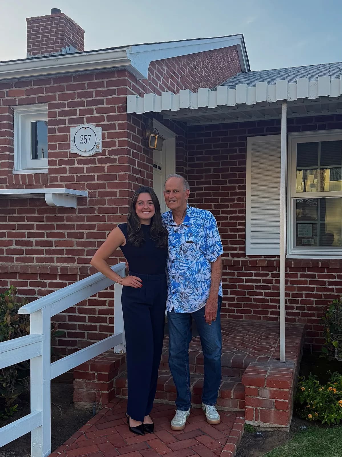 A younger woman in a navy sleeveless top and trousers standing with an older man in a tropical-print shirt on a brick walkway in front of a brick house numbered 257