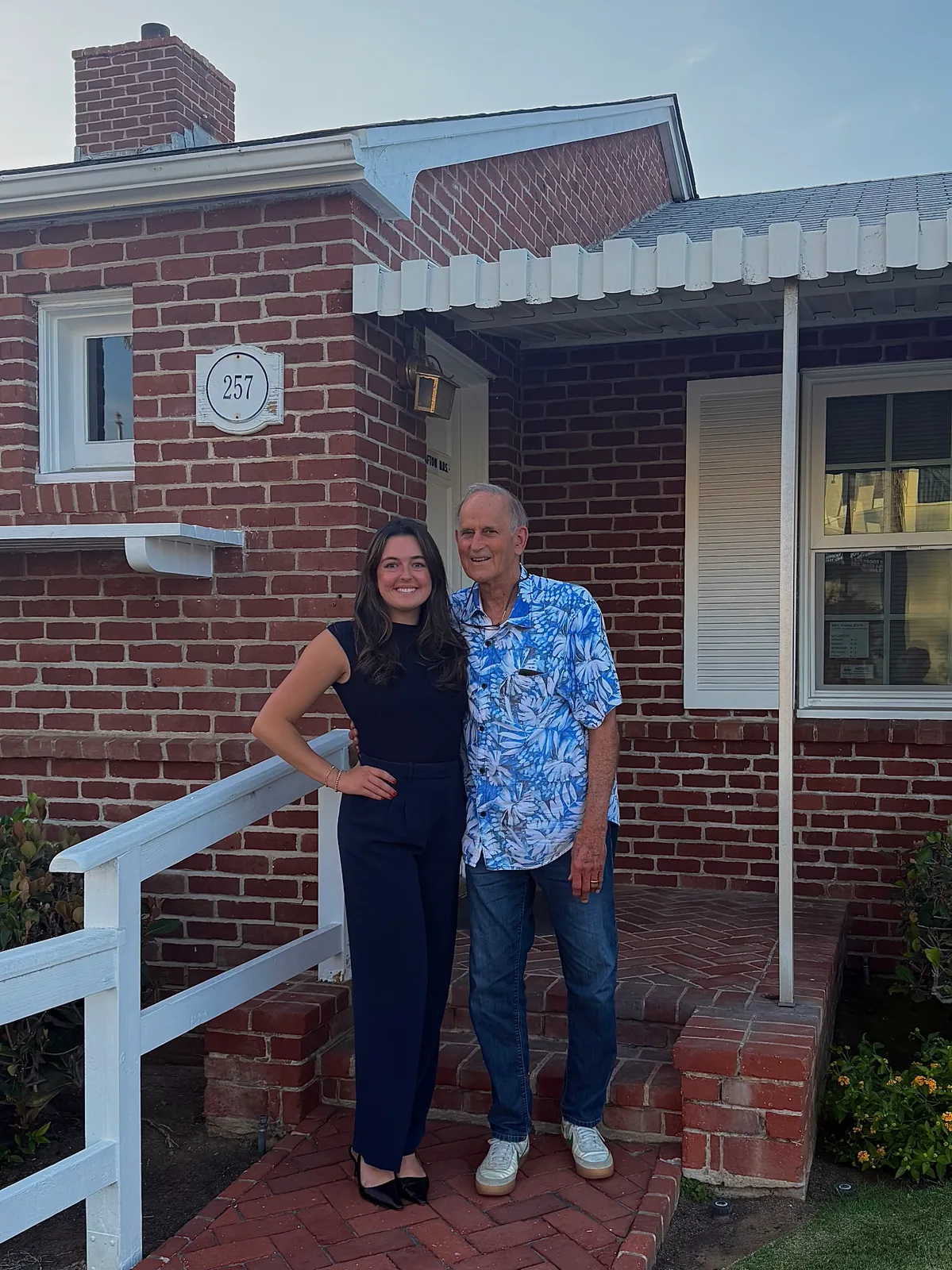 A younger woman in a navy sleeveless top and trousers standing with an older man in a tropical-print shirt on a brick walkway in front of a brick house numbered 257