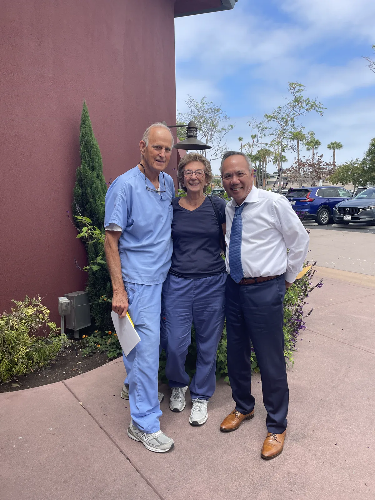 Three people standing outdoors: a man in light blue scrubs holding a yellow folder, a woman in navy scrubs, and a man in a white shirt and blue tie, in front of a burgundy wall
